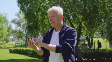 Yaşlı bir adam akıllı telefon kullanır ve parkta baş parmak işareti yapar. Beyaz saçlı emekliler internette fikir ve çözüm üretirler.
