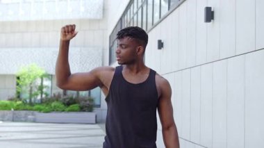 Spor giyim sektöründe Afro-Amerikalı bir adam bisepslerini gösteriyor ve kameraya gülümsüyor, atletik adam güç gösterisi ve kentsel geçmiş üzerine eğitim gösteriyor.