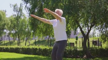 Beyaz tişörtlü atletik yaşlı adam yazın parkta egzersiz yapıyor ve gülümsüyor, aktif gri saçlı emekli spor yapıyor ve doğada aktif olarak rahatlıyor.