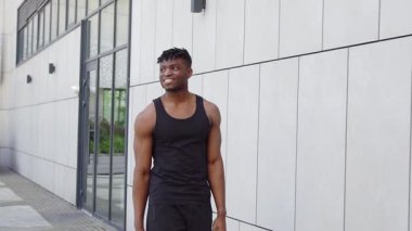 Spor kıyafetli Afrikalı bir adam duvarın arkasında yürüyor, atletik bir adam şehirde yürüyor ve gülümsüyor.