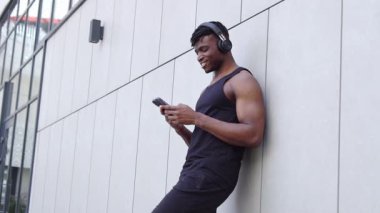 Afro-Amerikalı adam akıllı telefon kullanıyor ve sokakta müzik dinliyor, atletik adam cep telefonuyla yazı yazıyor ve şehir geçmişine gülümsüyor.