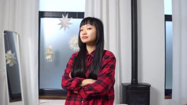 Asyalı kız kollarını kavuşturup evinde gülümsüyor, Koreli genç kadın saçını düzeltiyor ve göz kırpıyor.