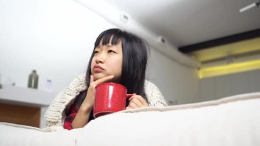 Asyalı düşünceli kadın kanepede oturup çay içerken düşünüyor, elinde fincanla hayal kuruyor ve evde hayal kuruyor.