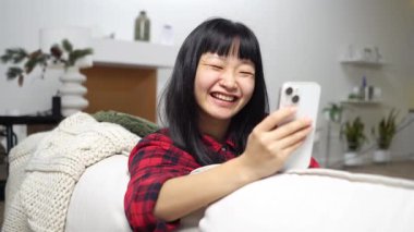 Asyalı genç bir kadın evde telefonla konuşuyor ve selamlıyor, Koreli bir kız kanepede görüntülü konuşuyor ve gülümsüyor.