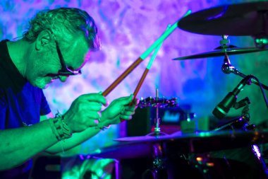 Yetişkin bir adamın gece bir konser sırasında davul çalıp gülümsemesi, yeşil, mavi ve pembe ışıklarla aydınlatılması. Canlı müzik kavramı.