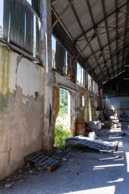 Çürüyen metal çatılı, yıpranmış beton duvarlı ve parlak doğal ışık altında etrafa saçılmış çöplü bir fabrika binasının yüksek açılı görüntüsü.