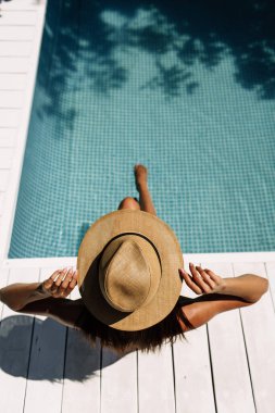 Bir kız bütün vücudu kaplayan hafif bir şapka tutuyor. Sırtı kameraya dönük bir şekilde havuzun kenarında oturuyor.