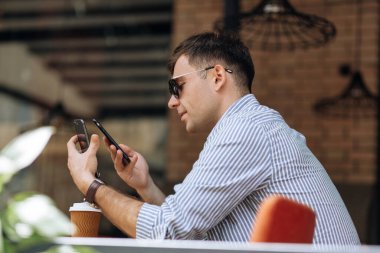 Bir kafede otururken telefona gülümseyen güneş gözlüklü yakışıklı esmer adam.