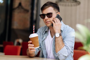 Güneş gözlüklü yakışıklı esmer adam bir kafede oturuyor ve telefonda gülümsüyor.
