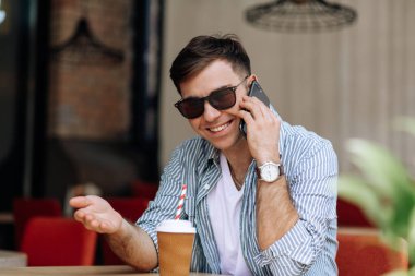 Güneş gözlüklü yakışıklı esmer adam bir kafede oturuyor ve telefonda gülümsüyor.