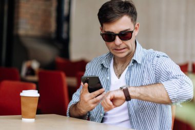 Güneş gözlüklü yakışıklı esmer adam kafede otururken kol saatine bakıyor.