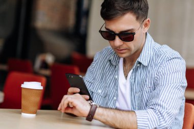 Güneş gözlüklü yakışıklı esmer adam kafede otururken kol saatine bakıyor.