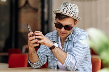 Şapkalı ve güneş gözlüklü yakışıklı bir adam bir kafede oturmuş elinde telefonla saatine bakıyor.