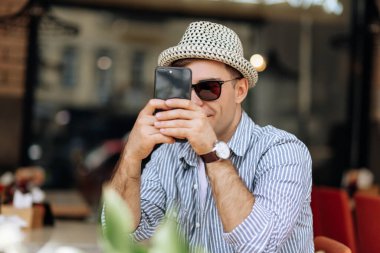 Güneş gözlüklü yakışıklı esmer adam ve elinde telefonla kafede gülümseyen bir şapka.