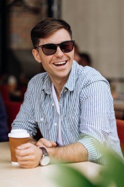 Güneş gözlüklü yakışıklı esmer adam sıcak bir yaz gününde bir kafede kahve içerken gülümsüyor.