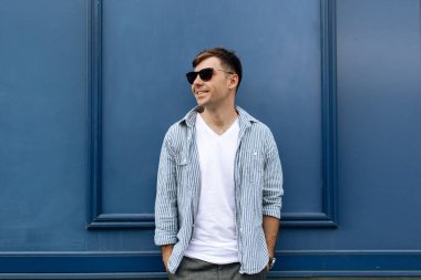 Yakışıklı, gülümseyen, güneş gözlüklü genç bir adamın portresi mavi arka planda eğleniyor.