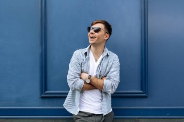 Yakışıklı, gülümseyen, güneş gözlüklü genç bir adamın portresi mavi arka planda eğleniyor.