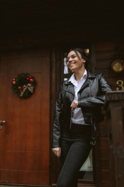 Deri ceketli esmer, ormandaki kahverengi ahşap bir evin verandasında gülüyor.