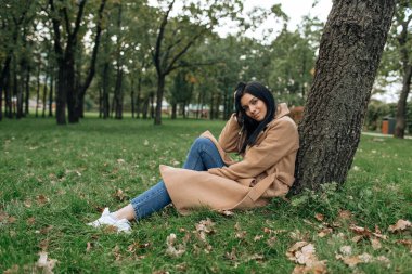 Kahverengi ceketli güzel esmer yeşil parktaki bir ağacın altında otururken gülümsüyor.