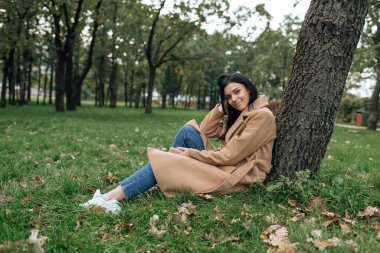 Kahverengi ceketli güzel esmer yeşil parktaki bir ağacın altında otururken gülümsüyor.