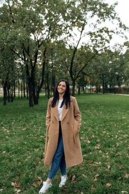 Kahverengi ceketli güzel esmer yeşil bir parkta yürüyor.