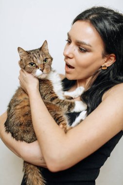 Güzel esmer kız şaşırmış yüzüyle bir kediyi kollarında tutuyor.