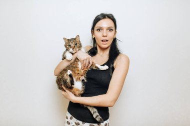 Güzel esmer kız şaşırmış yüzüyle bir kediyi kollarında tutuyor.