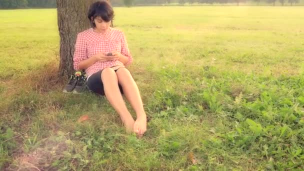 Chica enviando mensaje de texto bajo un árbol — Vídeos de Stock