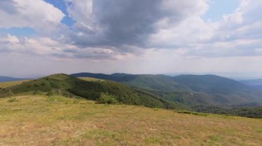Yuvarlanan yeşil tepeleri, bulutlu gökyüzünü ve güneş ışığını kullanarak barışçıl ve doğal bir atmosfer oluşturan manzaralı dağ manzarası..