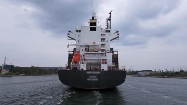 Bulutlu gökyüzünün altındaki limana giren büyük bir kargo gemisinin havadan arka görüntüsü. Deniz taşımacılığı ve küresel nakliye kavramı.