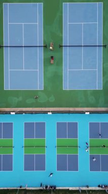 Parlak mavi ve yeşil yüzeylerde antrenman yapan oyuncuların bitişik tenis ve yankesicilik sahalarının havadan görünüşü yukarıdan canlı bir spor kompleksini gösteriyor..