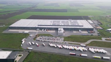 Yükleme rıhtımları ve kamyonları olan büyük bir lojistik deposunun havadan görünüşü, tarım arazileriyle çevrili dağıtım merkezi. Tedarik zinciri ve ulaşım kavramı.