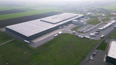 Yükleme rıhtımları ve kamyonları olan büyük bir lojistik deposunun havadan görünüşü, tarım arazileriyle çevrili dağıtım merkezi. Tedarik zinciri ve ulaşım kavramı.