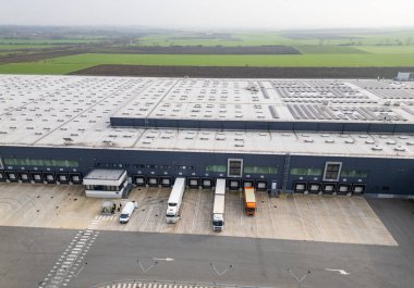 Yükleme rıhtımları ve kamyonları olan büyük bir lojistik deposunun havadan görünüşü, tarım arazileriyle çevrili dağıtım merkezi. Tedarik zinciri ve ulaşım kavramı.