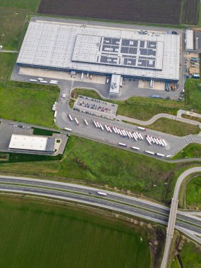 Yükleme rıhtımları ve kamyonları olan büyük bir lojistik deposunun havadan görünüşü, tarım arazileriyle çevrili dağıtım merkezi. Tedarik zinciri ve ulaşım kavramı.