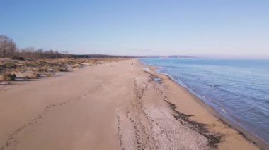 Boş kumsal ve berrak mavi gökyüzünün altında sakin deniz manzarası. Yumuşak dalgaları, kumulları ve uzak ufku olan huzurlu kıyı şeridi seyahat, doğa ve rahatlama temaları için ideal..