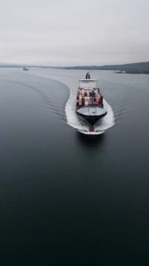 Sakin denizde ilerleyen büyük konteynır gemisinin havadan görünüşü ardında iz bırakıyor. Kargo gemisi, küresel ticaret, lojistik ve deniz sanayiinin sembolü olan kıyı şeridi boyunca yük taşımaktadır..