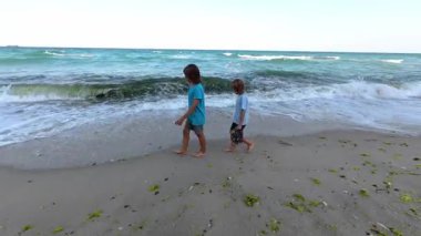Kumlu bir sahilde çıplak ayakla yürüyen iki çocuk, ellerinde ayakkabıları var. Yaz sahnesi dalgalar, mavi gökyüzü ve kaygısız çocukluk atmosferi, seyahat ve aile temaları için ideal.