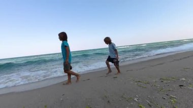 Kumlu bir sahilde çıplak ayakla yürüyen iki çocuk, ellerinde ayakkabıları var. Yaz sahnesi dalgalar, mavi gökyüzü ve kaygısız çocukluk atmosferi, seyahat ve aile temaları için ideal.