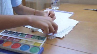 Kalem ve resimlerle dolu bir masada kağıda resim çizen bir çocuk. Yaratıcı etkinlik, öğrenme ve hayal gücü gelişimi, evde veya okulda sanat uygulaması, çocukluk eğitimi ve yaratıcılık kavramı.