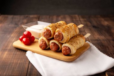 Yanında vişneli domates ve sosla birlikte susam tohumuyla puf böreğine sarılmış lezzetli sosis atıştırmalıkları. Ahşap tahtada servis edilir.