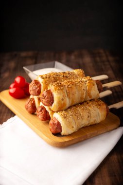 Çeri domates ve sosla birlikte susam tohumlarıyla puf böreğine sarılmış lezzetli sosisler. Ahşap tahtada servis edilir.