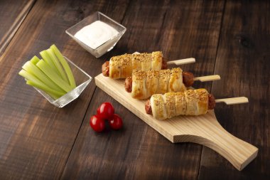 Çeri domatesleri, kereviz çubukları ve sos eşliğinde susam tohumlu puf böreğine sarılmış lezzetli sosisler. Ahşap tahtada servis edilir.