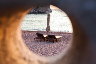 Sahil sahnesi, kumlu sahilde iki güneş paneli ve kapalı şemsiyeyi gösteren yuvarlak bir açıklıktan çerçevelenmiş, sakin, samimi bir tatil atmosferi yaratıyor..