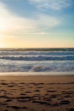 Sahilde gün batımında parlayan gökyüzü, kıyıya vuran dalgalar ve dalgalarda tek bir karanlık kaya, sakin ama dramatik bir kıyı havası yaratır..