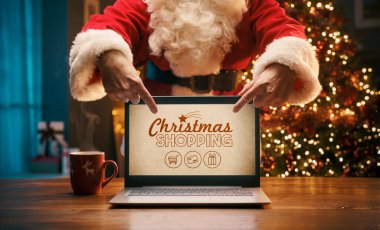 Noel Baba dizüstü bilgisayarında Noel reklamları gösteriyor. Bilgisayar ekranını işaret ediyor.