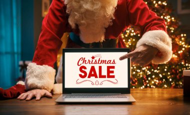 Noel Baba dizüstü bilgisayarında Noel reklamları gösteriyor. Bilgisayar ekranını işaret ediyor.