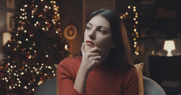 Lonely woman thinking with hand on chin at Christmas, tree with lights in the background