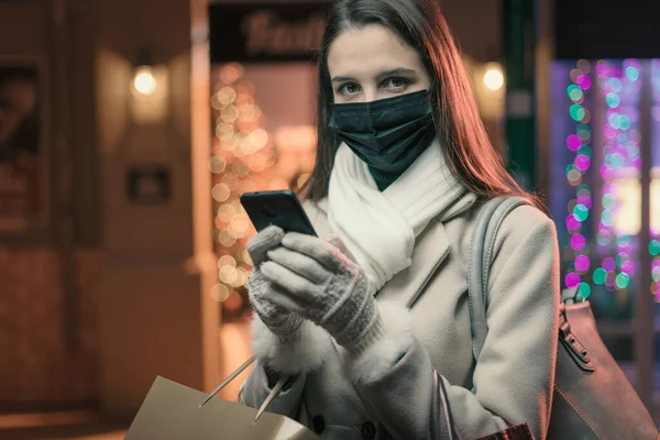 女性はクリスマスの買い物をし コロナウイルスの感染を防ぐために保護顔マスクを着て 彼女は彼女のスマートフォンを使用しています — ストック写真