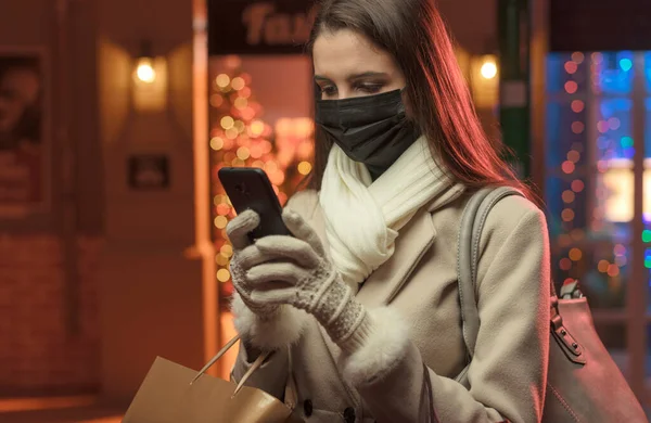 女性はクリスマスの買い物をし コロナウイルスの感染を防ぐために保護顔マスクを着て 彼女は彼女のスマートフォンを使用しています — ストック写真